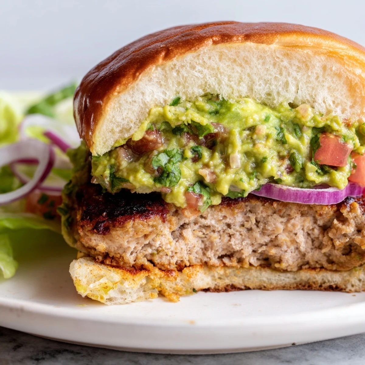 Golden-brown turkey burger topped with creamy guacamole, lettuce, and red onion on a toasted bun served with sweet potato fries.
