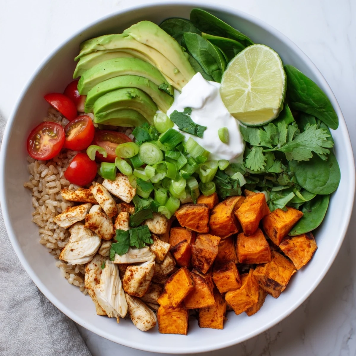 Savory slices of chicken and caramelized sweet potatoes rest on brown rice, topped with spinach, cherry tomatoes, cilantro, and lime in this bowl.